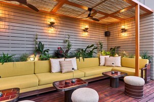 Uma área externa moderna com assentos, um sofá amarelo, mesas redondas e plantas em vasos.