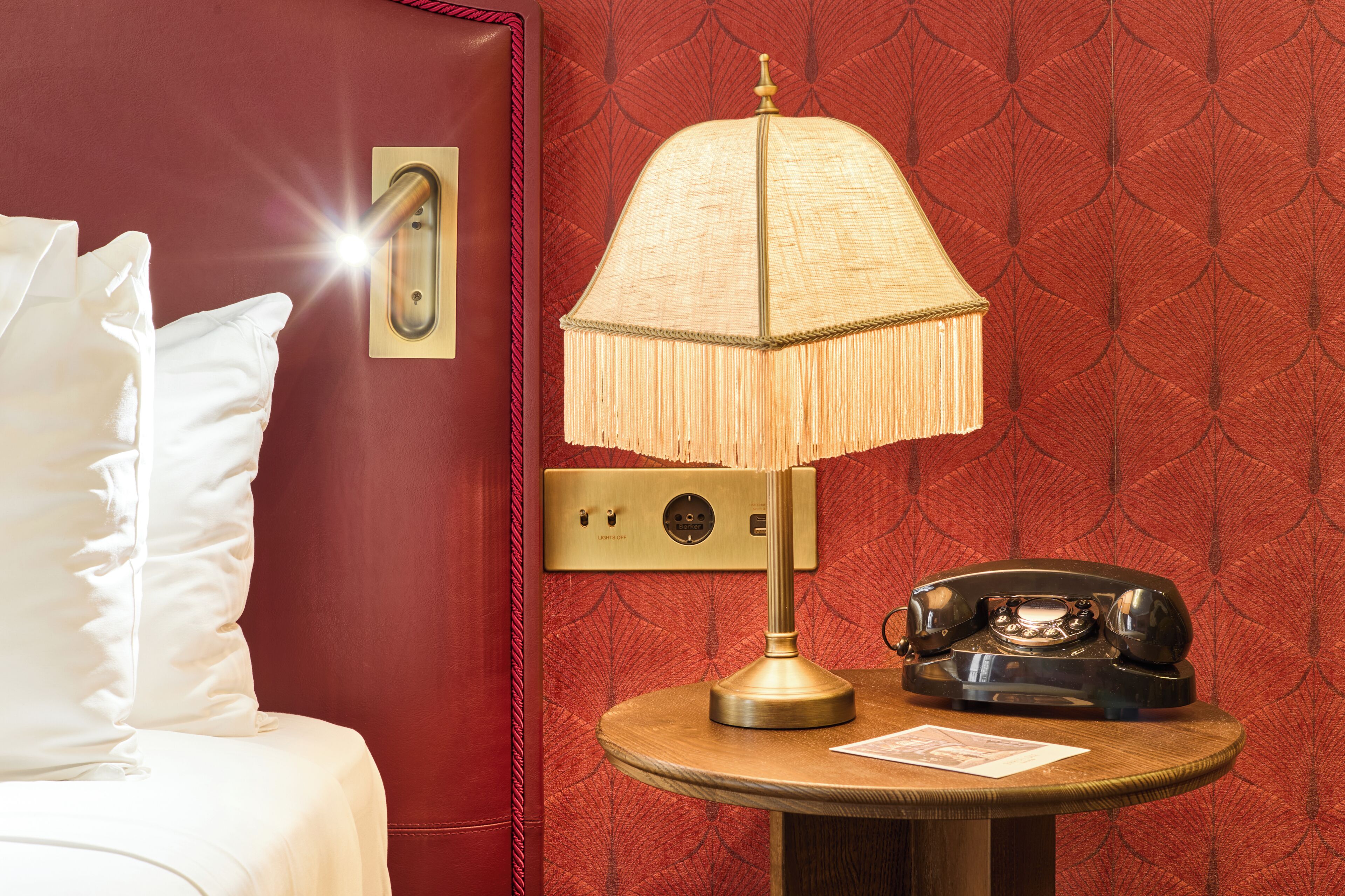Quarto de hotel com parede vermelha, mesa de madeira redonda com abajur, telefone antigo e travesseiro branco.