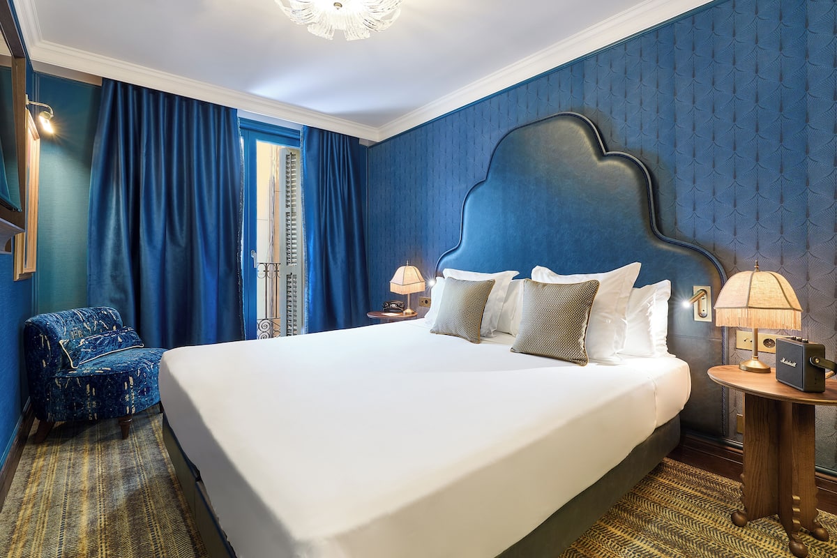 A hotel room with a large bed, a chair, a nightstand with a lamp, and a window with curtains.