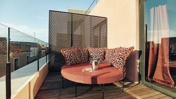 Mesa redonda con cojines estampados sobre una terraza de madera, una silla de estructura metálica y una partición de vidrio con diseño de malla.
