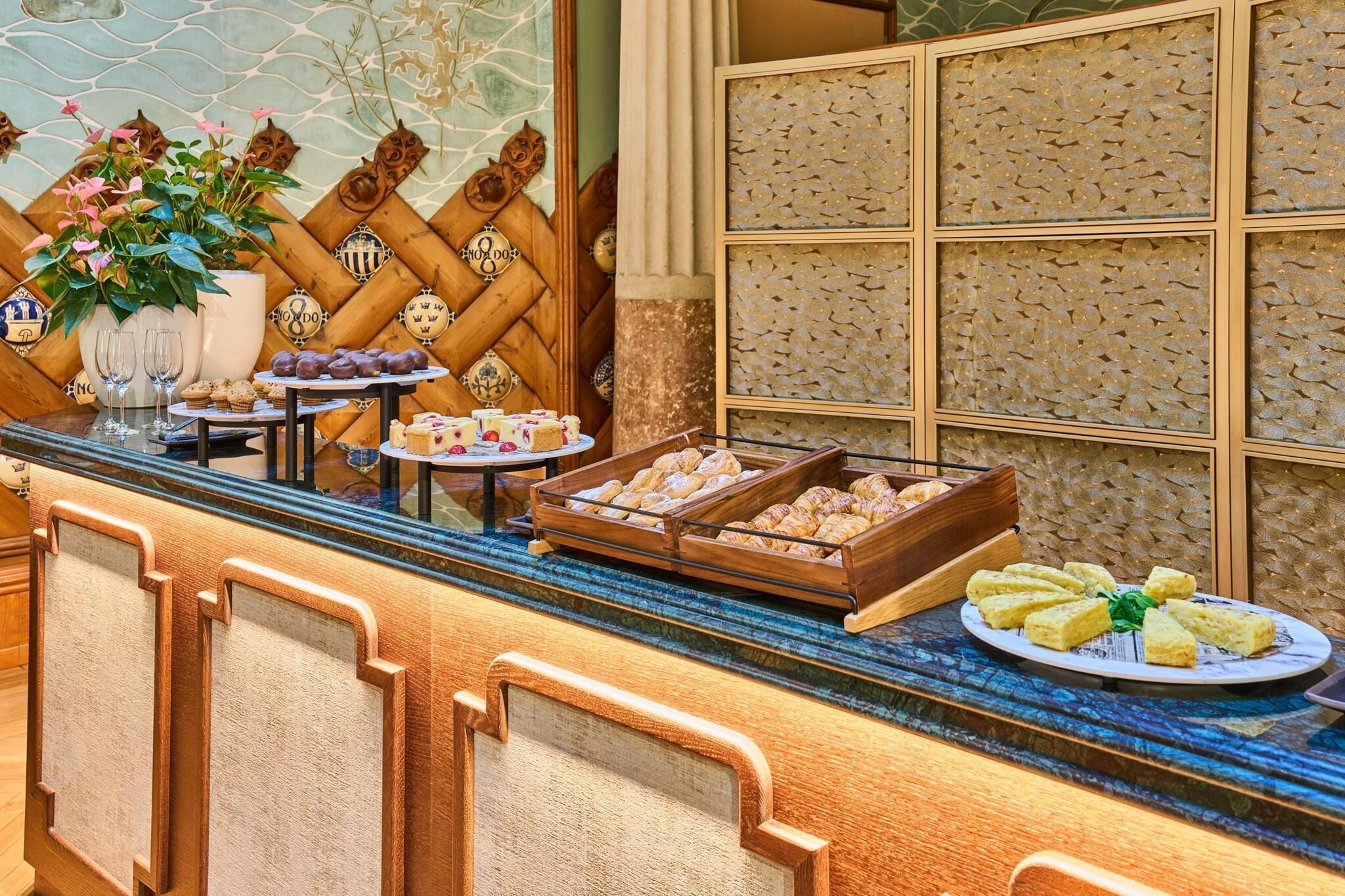 Un mostrador de buffet con diversos alimentos, entre ellos pasteles y postres, y una pared decorativa con un diseño geométrico.