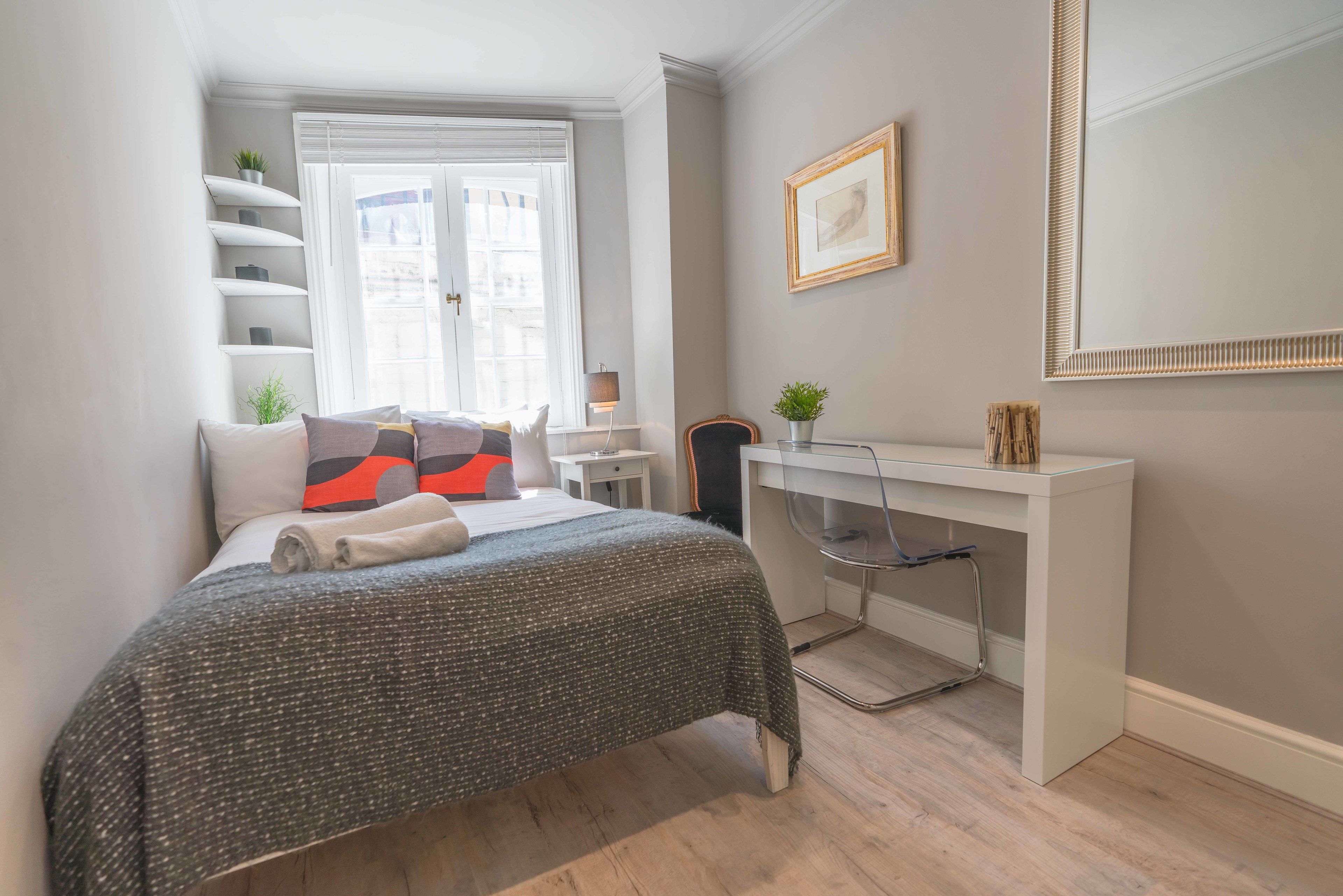 A bedroom with a bed, a desk, a chair, a window, and a mirror.