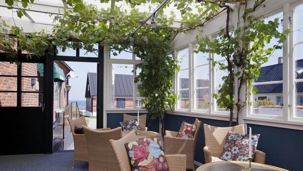 A cozy room with wicker furniture, a round table, and large windows with views of buildings. - Branteviks Bed & Breakfast (Brantevik)