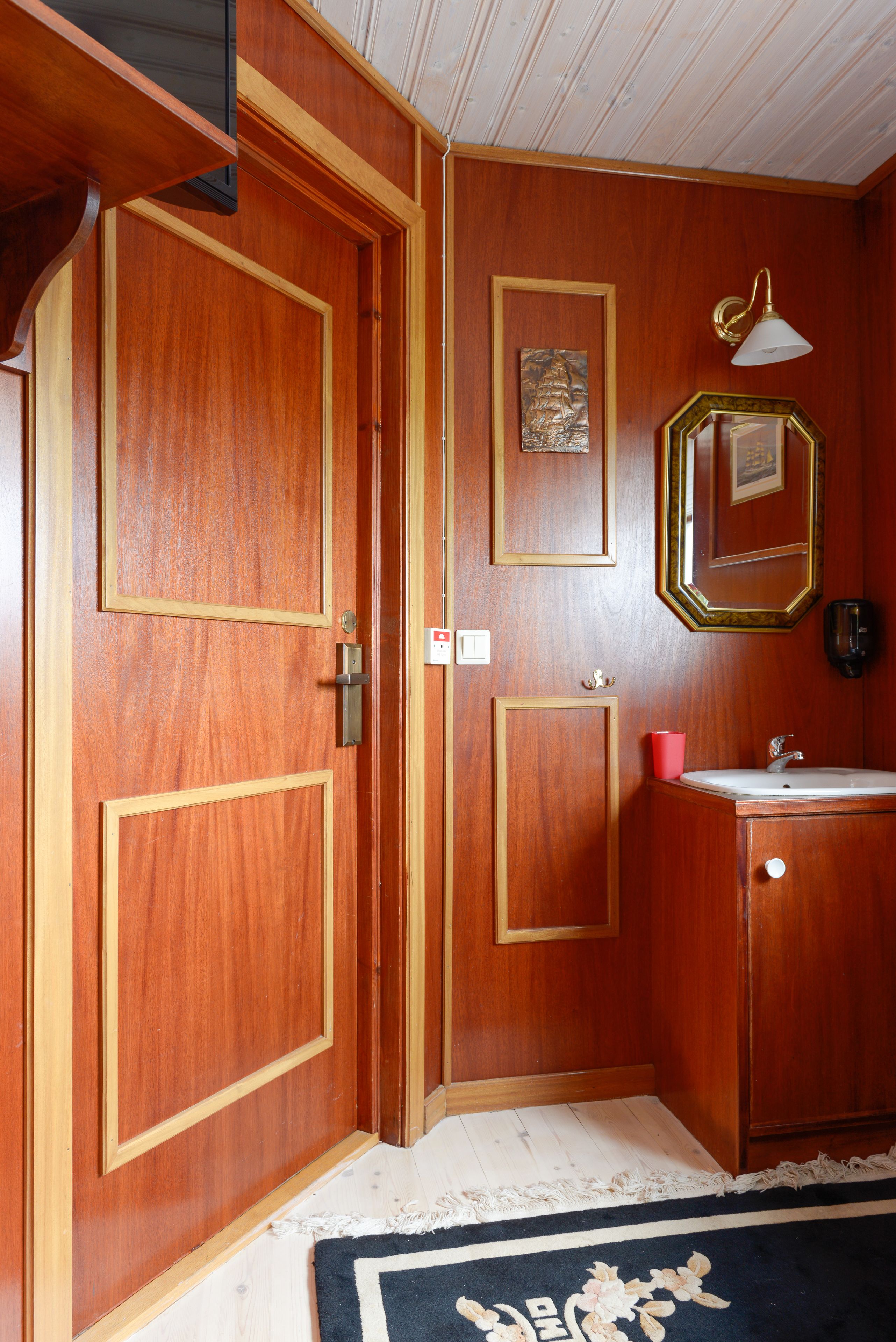 Una habitación con paneles de madera, una puerta con manija, un cuadro enmarcado en la pared, una lámpara y un lavabo con espejo.