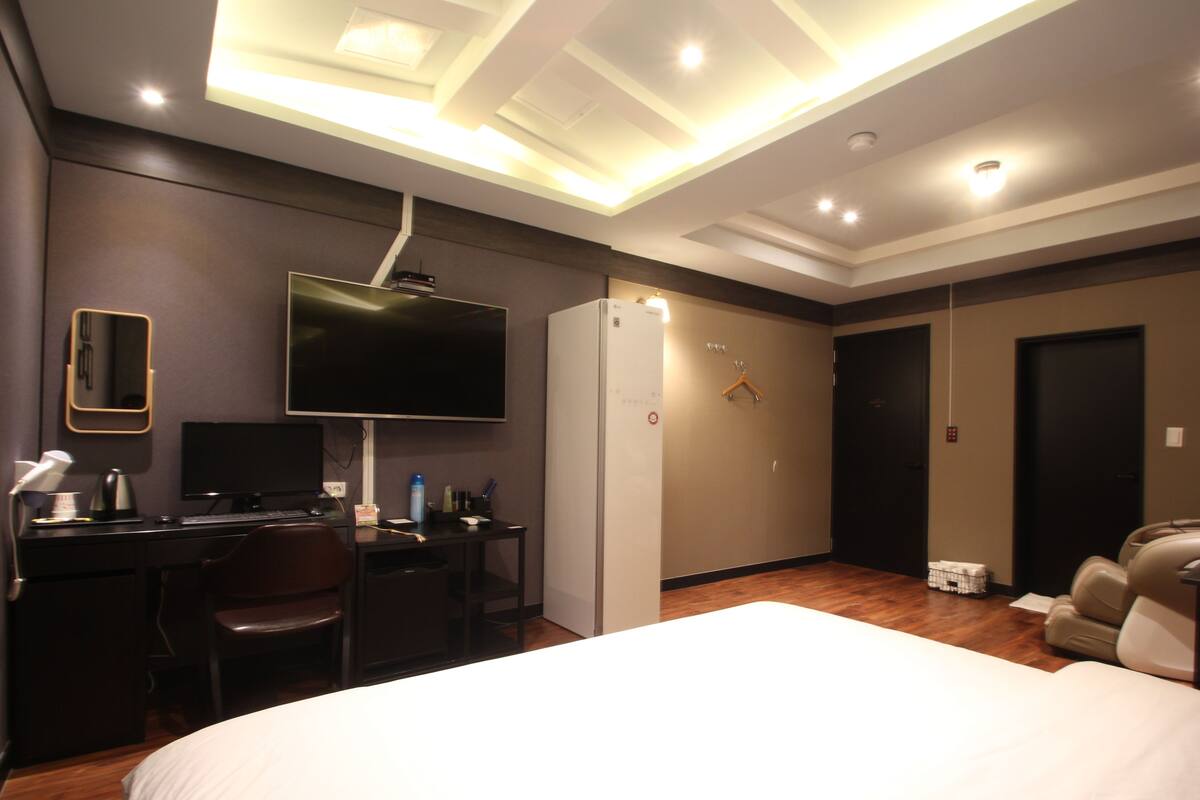 A hotel room with a bed, desk, chair, TV, and a massage chair.