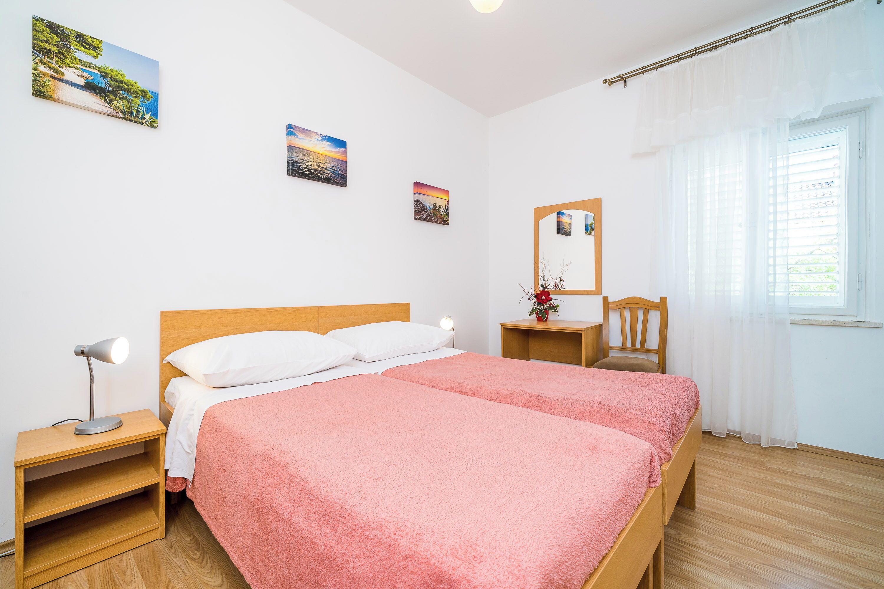 A double bed with pink bedding, a wooden nightstand with a lamp, a wooden chair, and a small table with a vase.