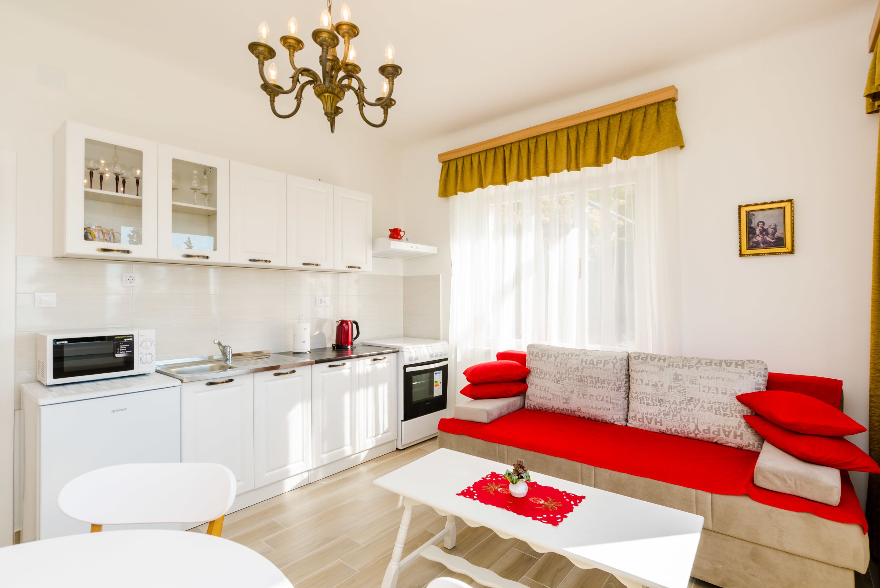 Une cuisine moderne avec des meubles blancs, un four à micro-ondes et une bouilloire rouge. On y trouve une table à manger blanche avec un set de table rouge et un vase. Un canapé rouge avec des coussins à motifs est placé contre un mur où est accroché un tableau encadré.