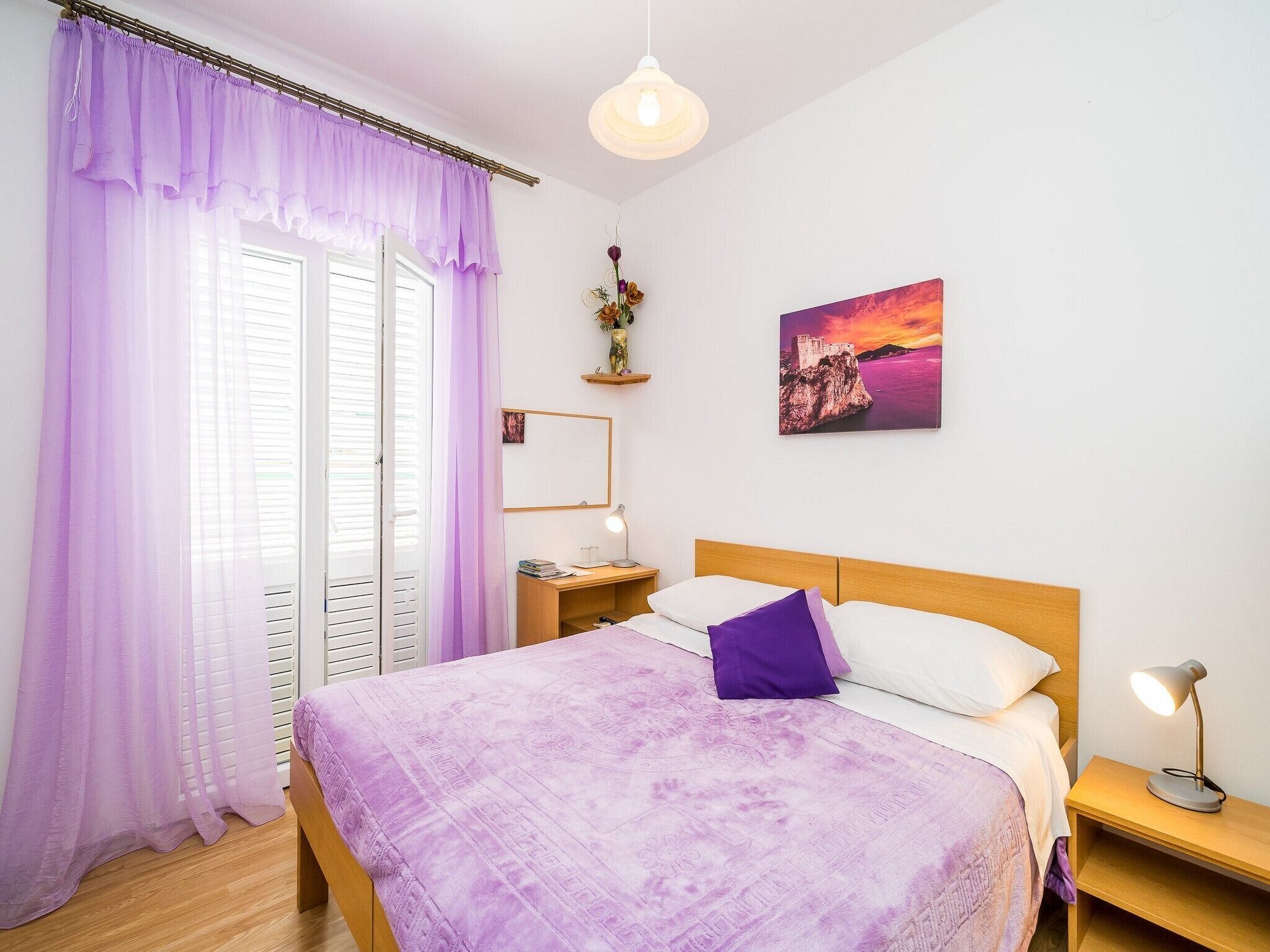 A bedroom with a bed, a nightstand, a desk, a chair, a window with curtains, a wall-mounted mirror, a painting, and a hanging light fixture.