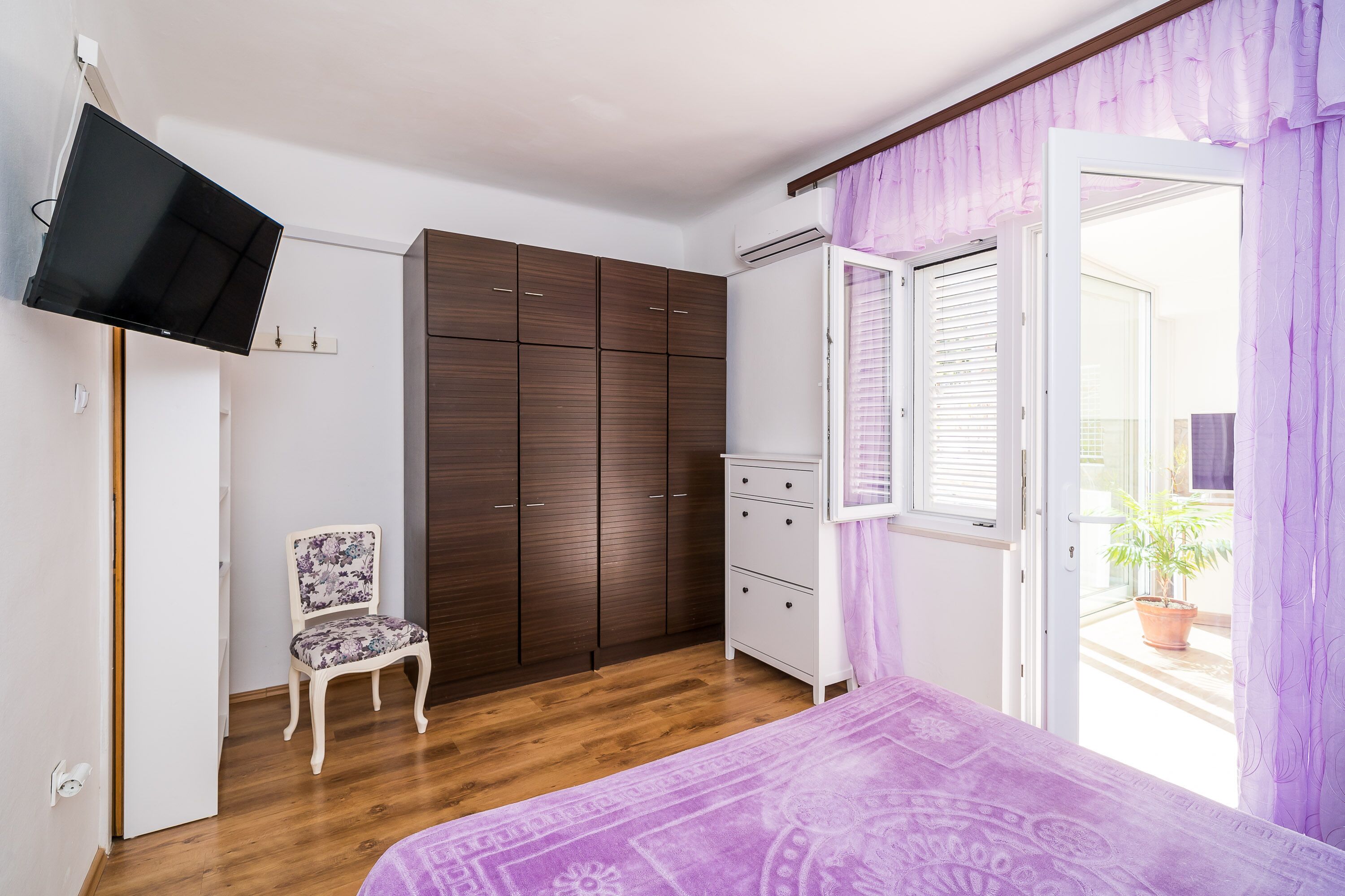 Een slaapkamer met een bed, een kledingkast, een stoel, een televisie en een raam met zonwering.