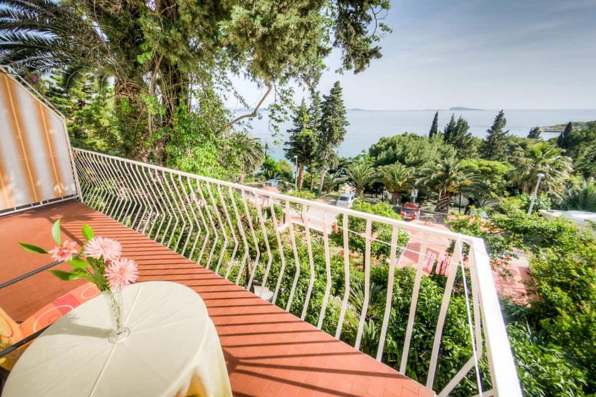 Een balkon met een witte tafel en stoelen, omringd door weelderig groen en uitzicht op zee.