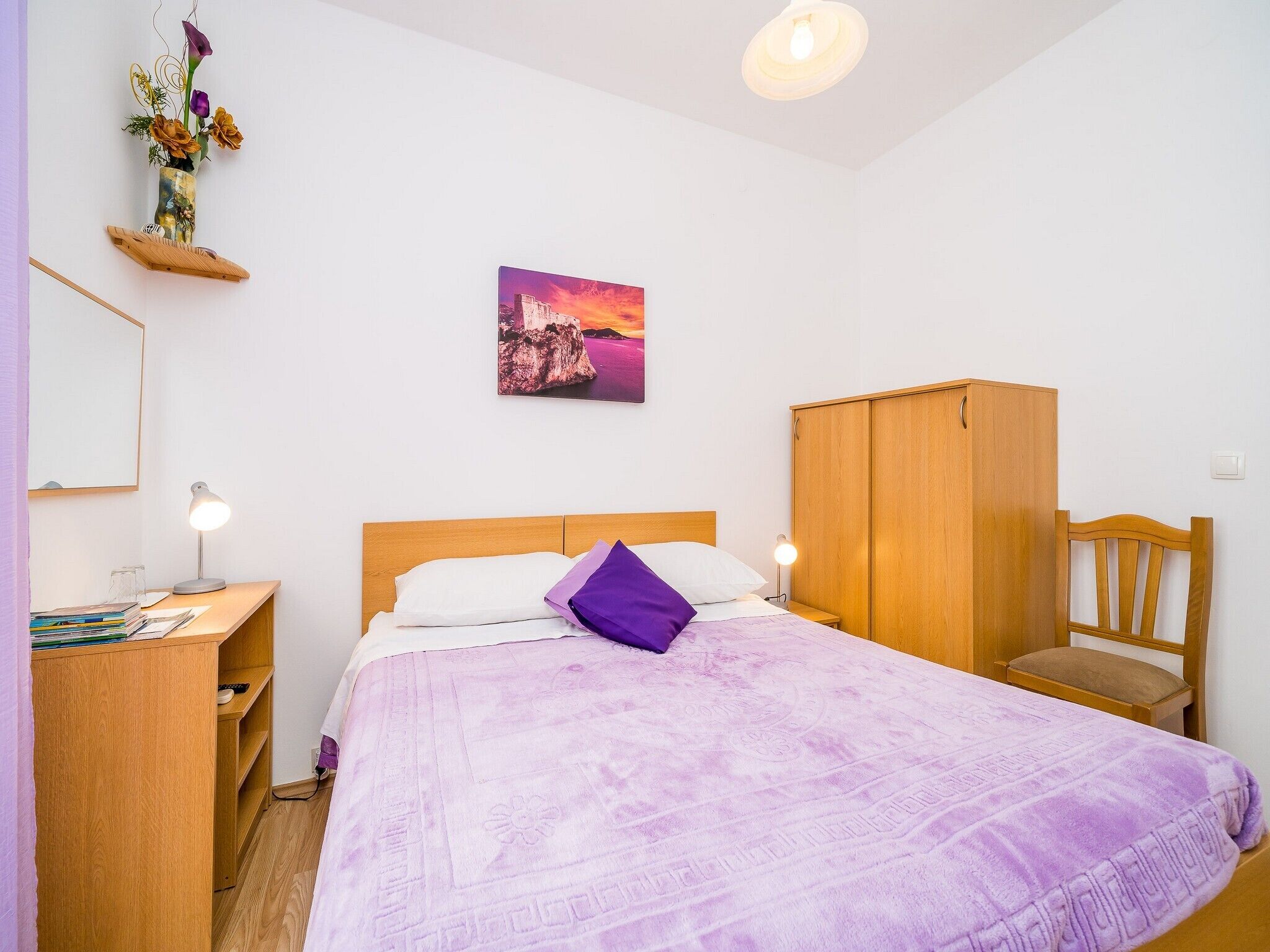 Une chambre à coucher avec un lit, une armoire en bois, un bureau avec une lampe et un tableau accroché au mur.
