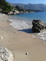 A beach with pebbles and a large rock in the foreground, a clear blue sea, and mountains in the background.