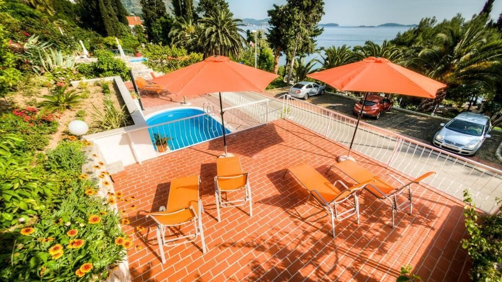 Un área de piscina en la azotea con sillas y sombrillas naranjas, rodeada de exuberante vegetación y vistas al mar.