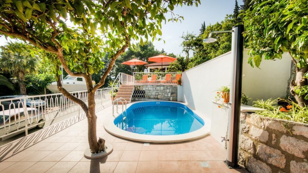 A swimming pool with a stone wall, a covered seating area with umbrellas, and a tree in the foreground.