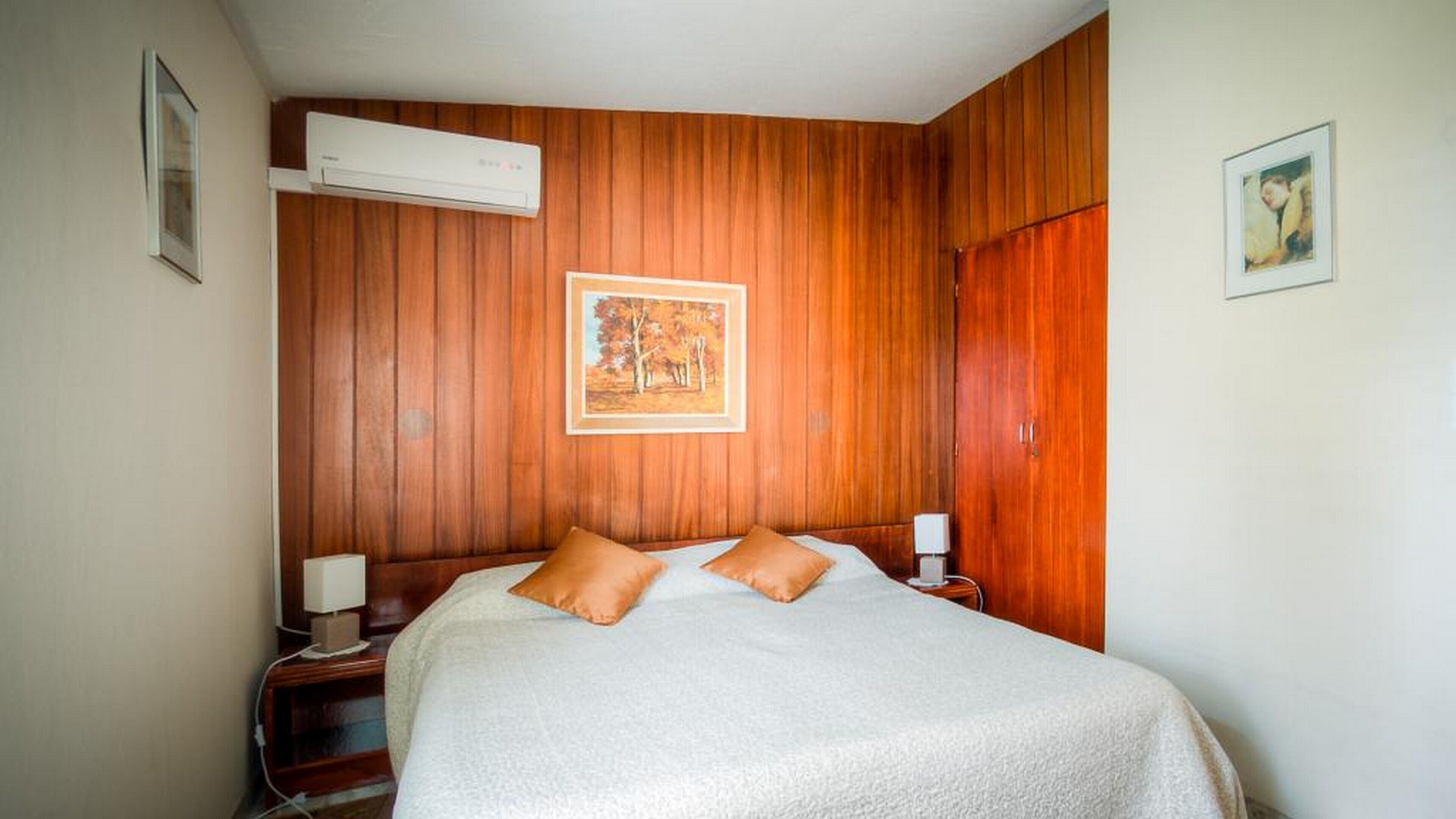 A hotel room with a wooden paneled wall, a bed with two pillows, a nightstand with a lamp, and an air conditioning unit.