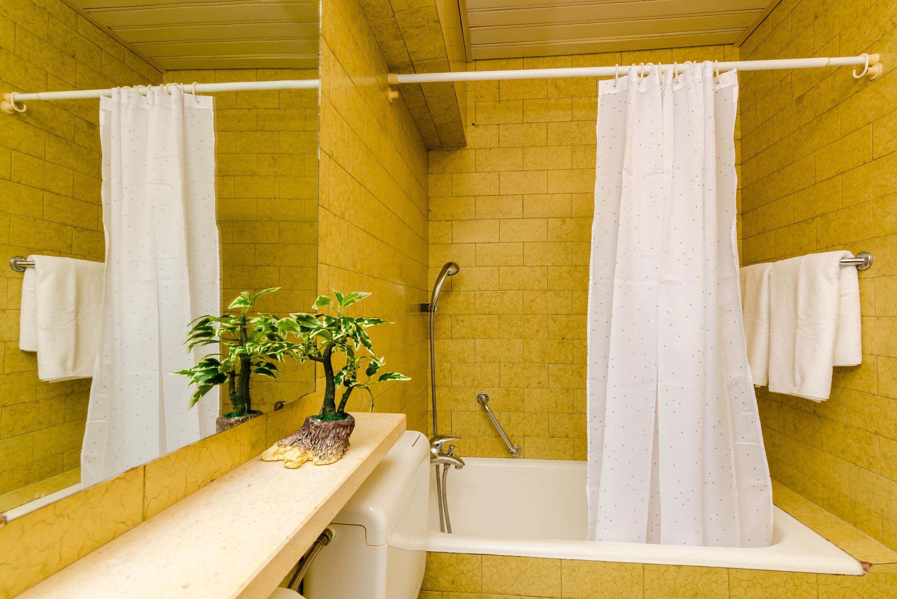 Ein Badezimmer mit gelben Fliesen, einer weißen Badewanne, einer Dusche mit weißem Vorhang und einem kleinen Topfpflanzen auf dem Ablagetisch.
