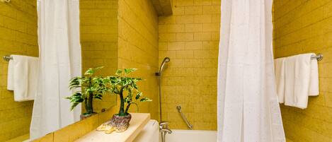 Un baño con paredes de azulejos amarillos, una bañera blanca, una ducha con cortina blanca y una pequeña planta en maceta sobre el mostrador.