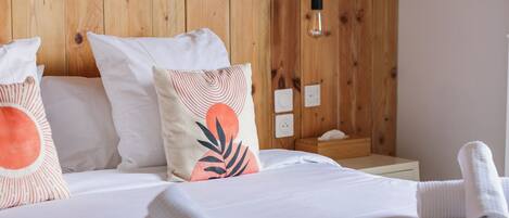 A neatly made bed with white linens and a rolled towel, a wooden headboard, and a wall-mounted light fixture.