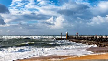 A coastal scene with a pier extending into the sea, featuring two lighthouses and a rocky shoreline.