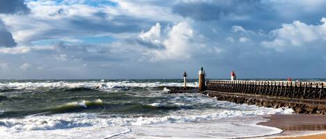 A coastal scene with a pier extending into the sea, featuring two lighthouses and a rocky shoreline.