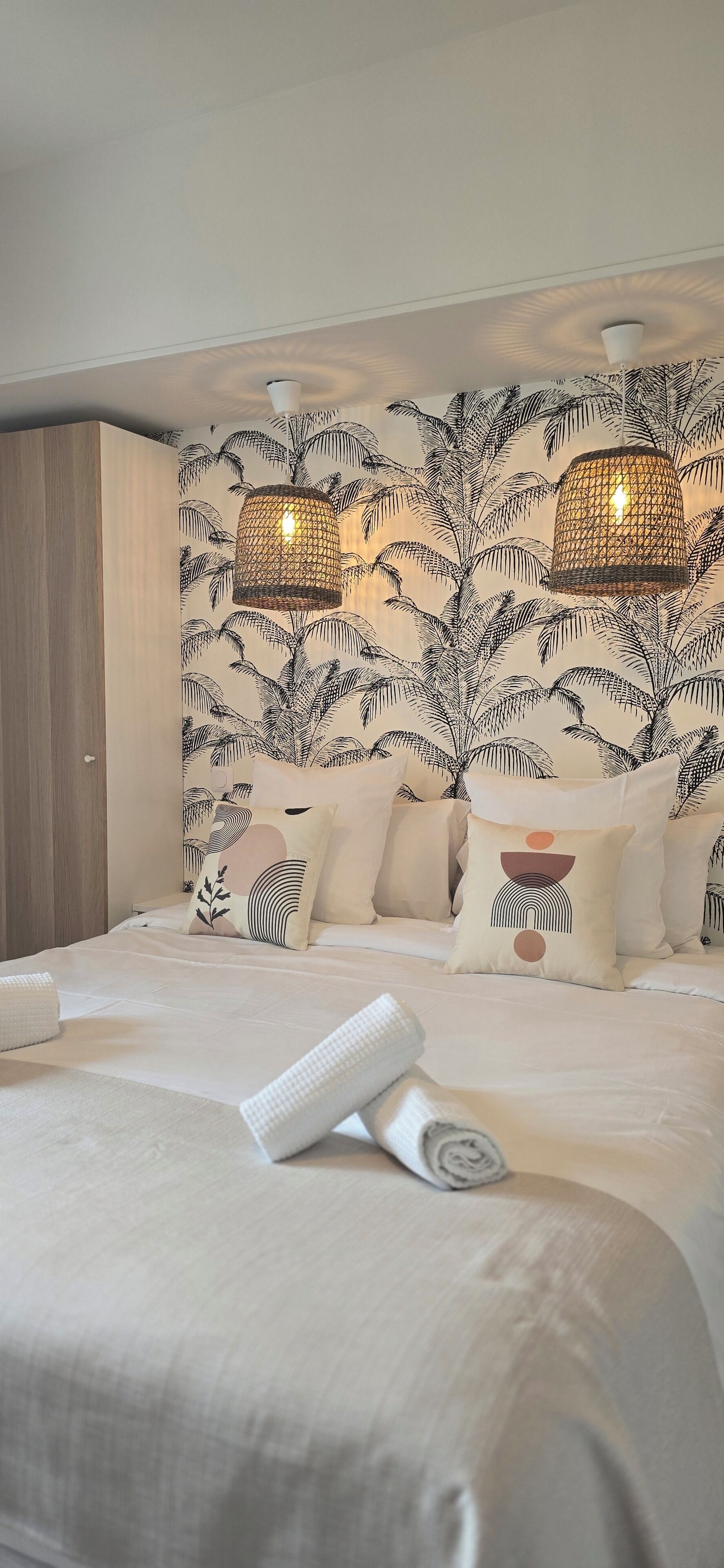 A neatly made bed with a rolled towel, a patterned wall, and two hanging lamps.