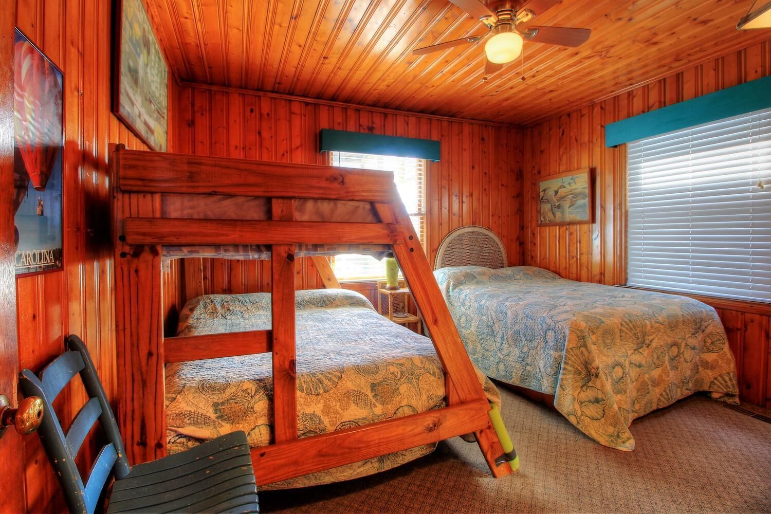 Une cabine avec deux lits, un lit superposé, des murs en bois, un ventilateur de plafond et une fenêtre avec des stores.