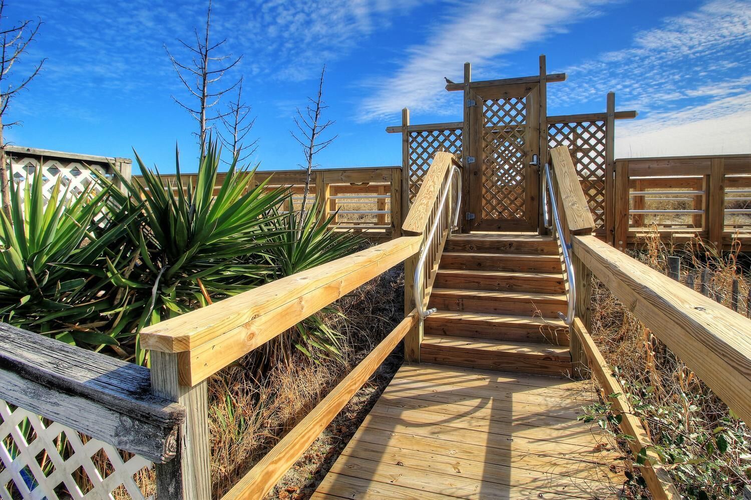 Eine Holztreppe führt zu einem verschlossenen Eingang, umgeben von trockener Vegetation und blauem Himmel.