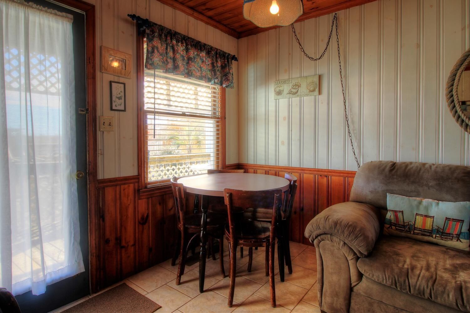 A cozy room with a round wooden table and chairs, a cushioned sofa with a beach-themed pillow, and a window with blinds.