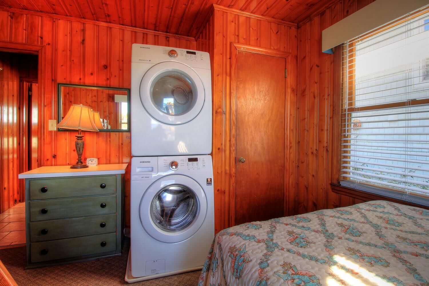 A room with a washing machine, a dresser, a lamp, and a bed.
