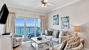 Un salon spacieux avec un balcon, un ventilateur de plafond, un téléviseur à écran plat et un tableau représentant une scène de plage.