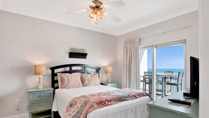 Une chambre à coucher comprenant un lit, deux tables de chevet, un ventilateur de plafond, une télévision et un balcon avec vue sur l’océan.