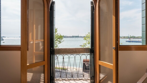Un balcón con vista a un río y barcos, un marco de puerta de madera y una barandilla de metal.