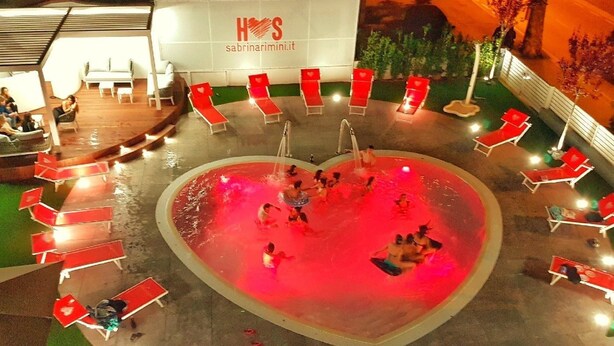 Ein herzförmiger Pool mit roter Beleuchtung, umgeben von roten Liegestühlen, und ein Schild mit der Aufschrift 'HOS'.