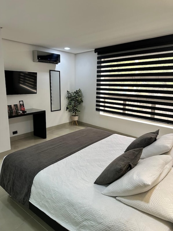 Une chambre à coucher moderne avec un lit, un téléviseur fixé au mur, une plante en pot et une fenêtre avec des stores.