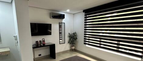 A modern bedroom with a large bed, a TV mounted on the wall, a desk, and a window with blinds.
