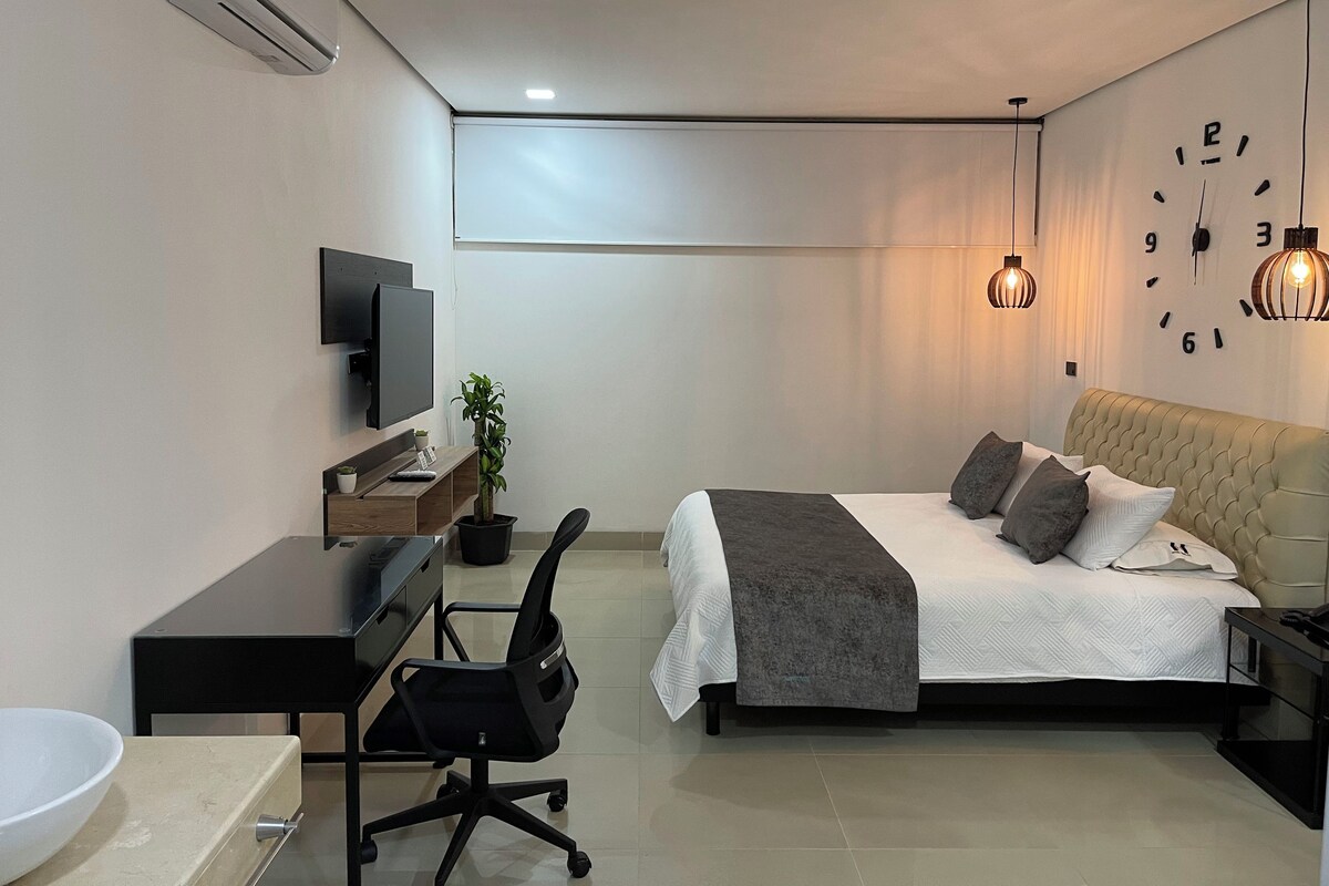 A modern bedroom with a bed, bedside table, lamp, TV, desk, chair, and a potted plant.