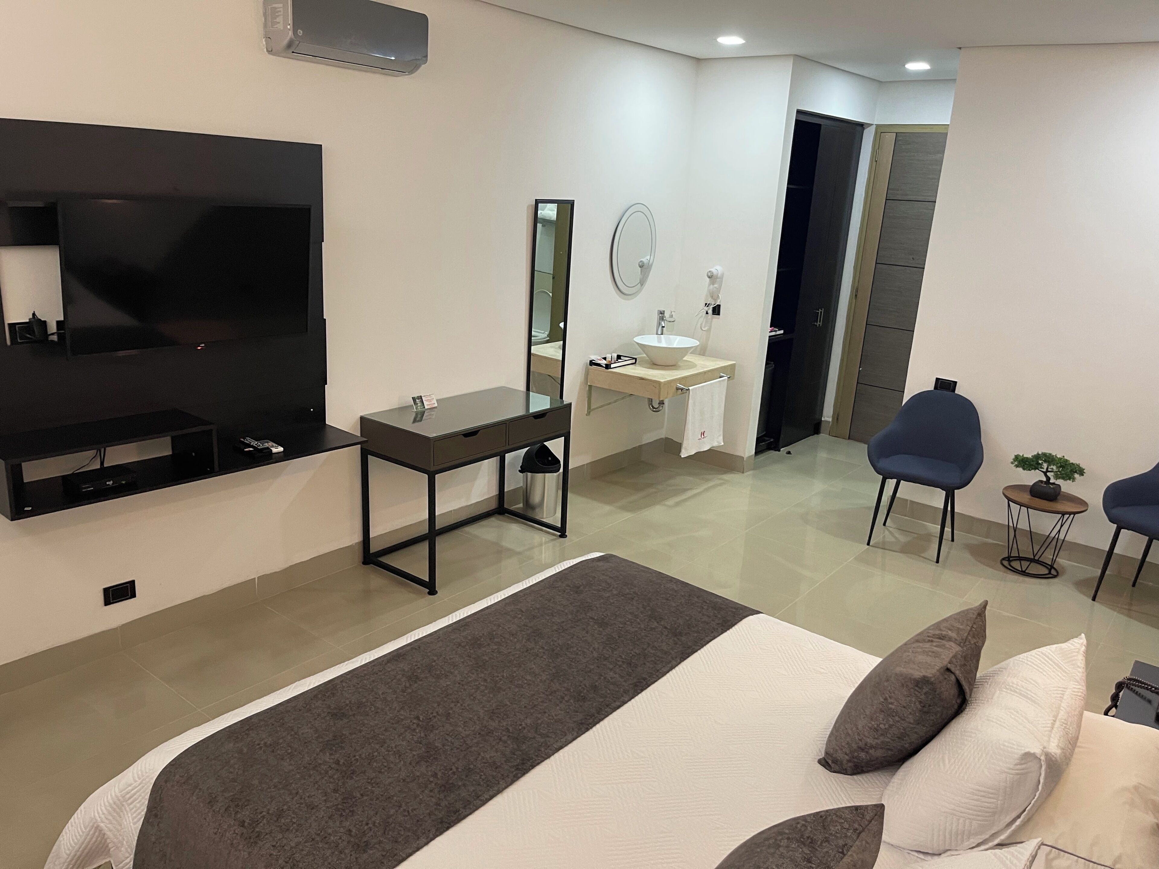 Quarto de hotel moderno com uma cama grande, televisão, escrivaninha com pia, espelho e duas cadeiras.