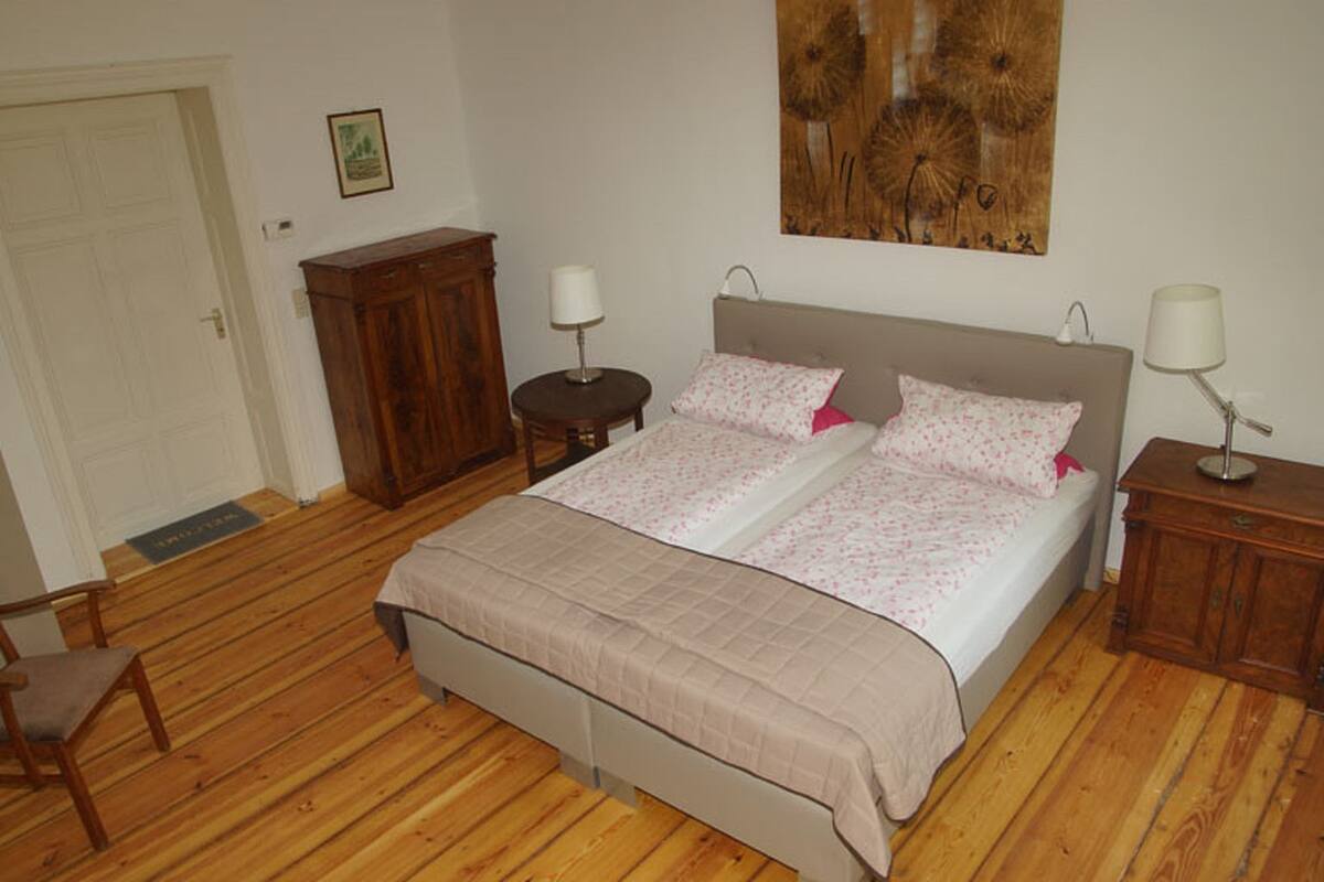 Un dormitorio con una cama doble, dos mesitas de noche, un armario de madera, una puerta y una obra de arte colgada en la pared.