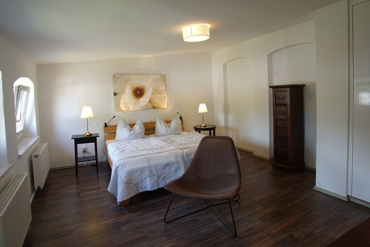 Un dormitorio con una cama, dos mesitas de noche con lámparas, una silla, un armario y un ventanal grande.