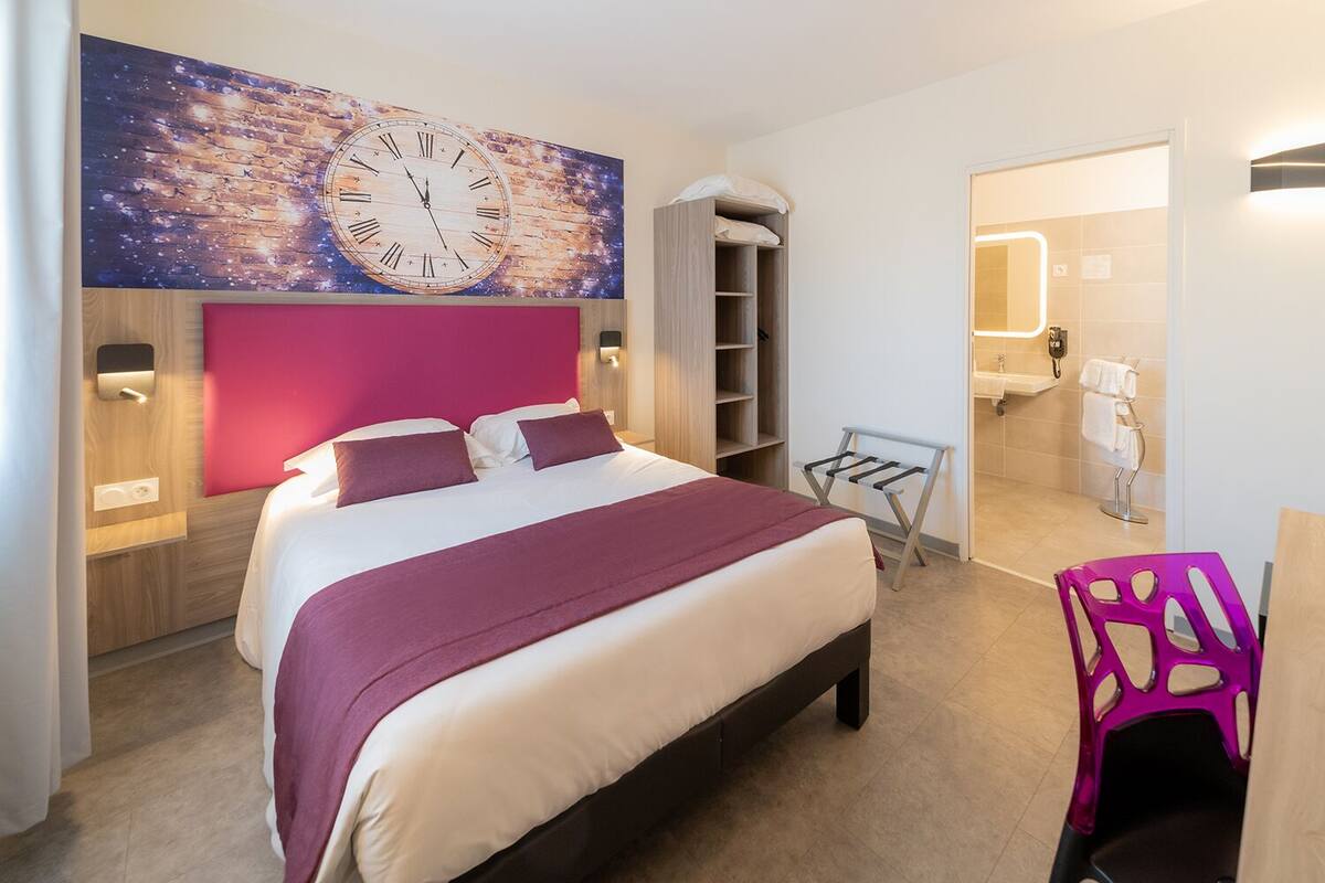Une chambre d’hôtel moderne dotée d’un grand lit, d’une horloge murale, d’une armoire, d’un petit coin salon et d’une salle de bain visible à travers une porte ouverte.