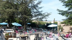 Un espace extérieur aménagé en terrasse, avec plusieurs tables et chaises, dont certaines sont placées sous des parasols bleus, le tout entouré d’arbres.
