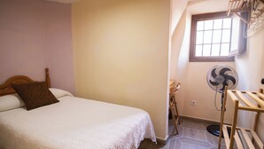 Un dormitorio con cama, cabecera de madera, ventana, ventilador de pie y estante de madera.