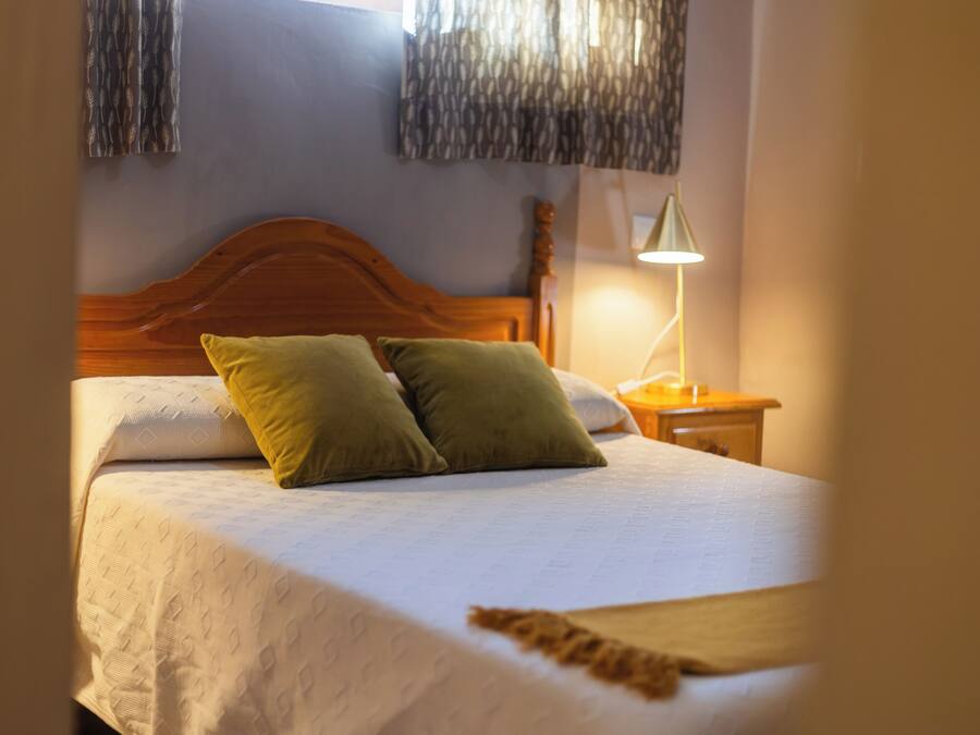 Una cama con cabecera de madera, dos almohadas, una lámpara de noche y una colcha estampada.