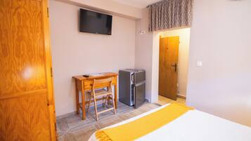 Una habitación con cama, escritorio con silla, televisor montado en la pared y refrigerador.