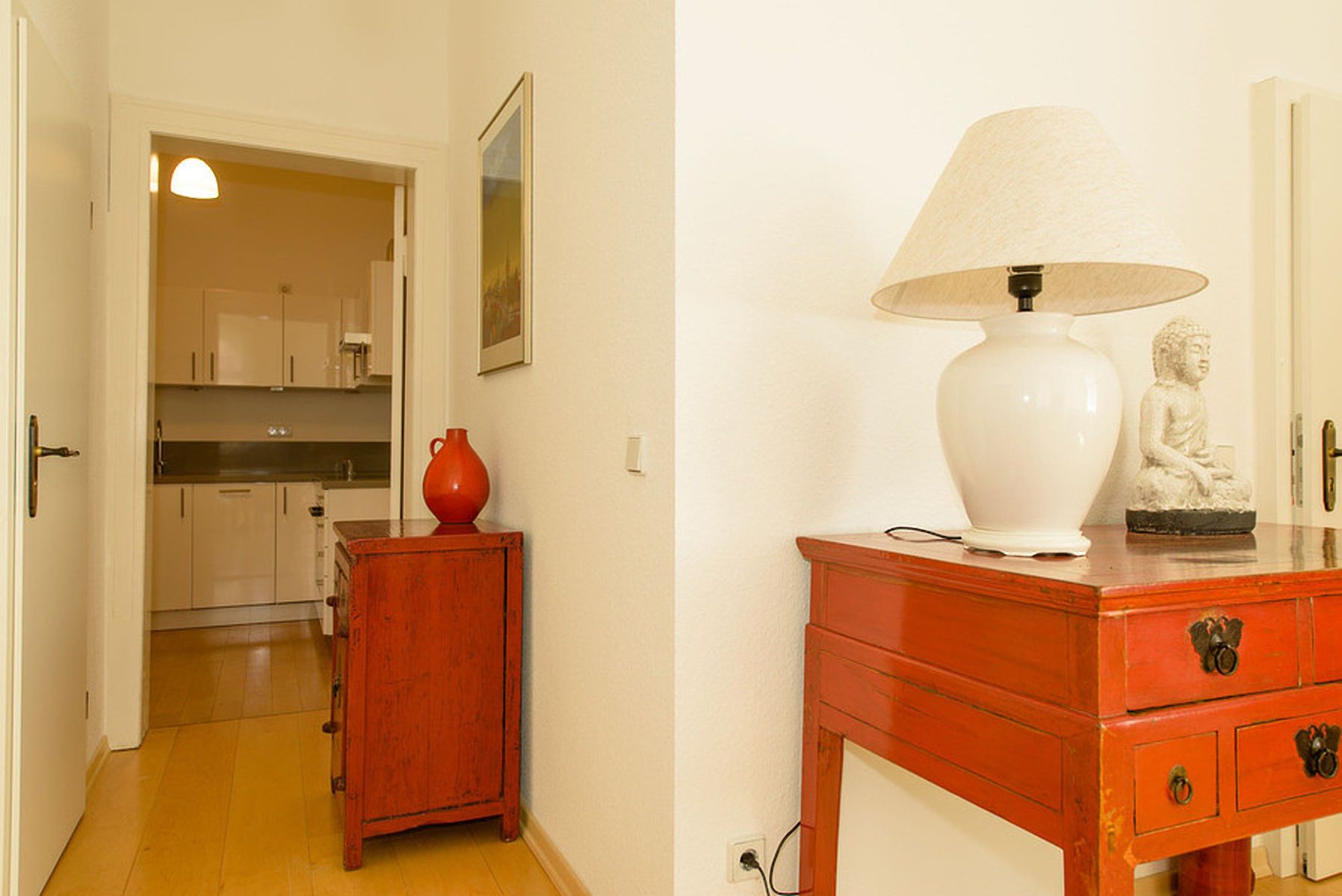 A hallway with a wooden cabinet, a red vase, a white lamp, and a statue on a wooden table.