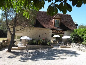 A stone building with a tiled roof, outdoor seating with umbrellas, and a gravel driveway. - Bergerie de Lagarrigue (Baladou)