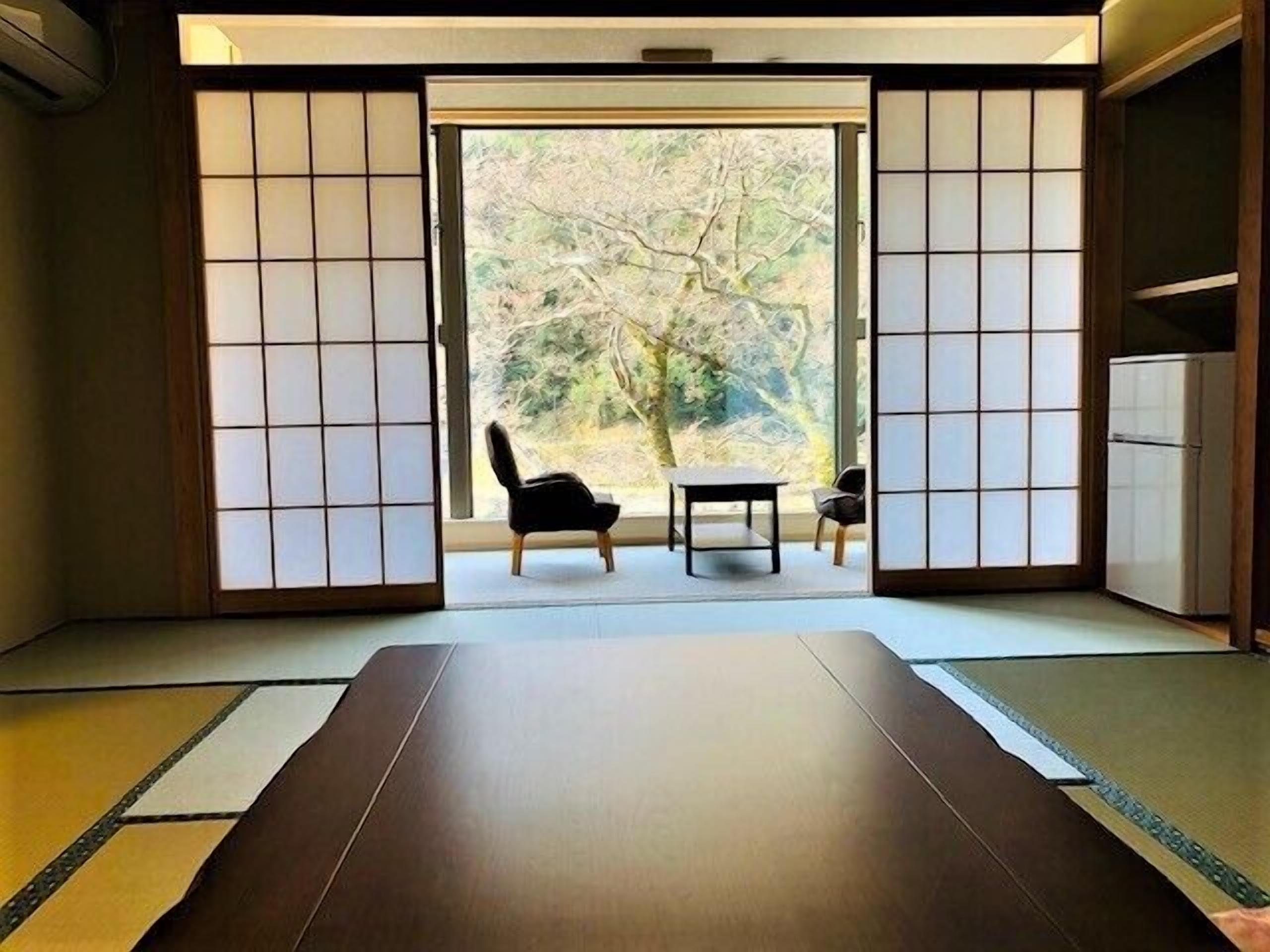 A traditional Japanese room with tatami flooring, a low table, two chairs, and a sliding door leading to a view of trees.