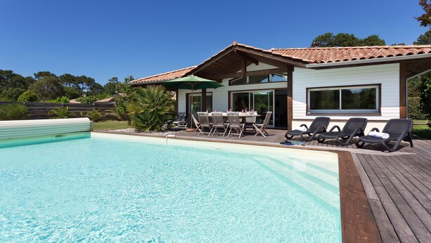 Une piscine avec une terrasse en bois, un patio couvert avec des chaises et une maison avec un toit en tuiles.