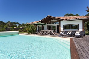 Une piscine avec une terrasse en bois, un patio couvert avec des chaises et une maison avec un toit en tuiles.