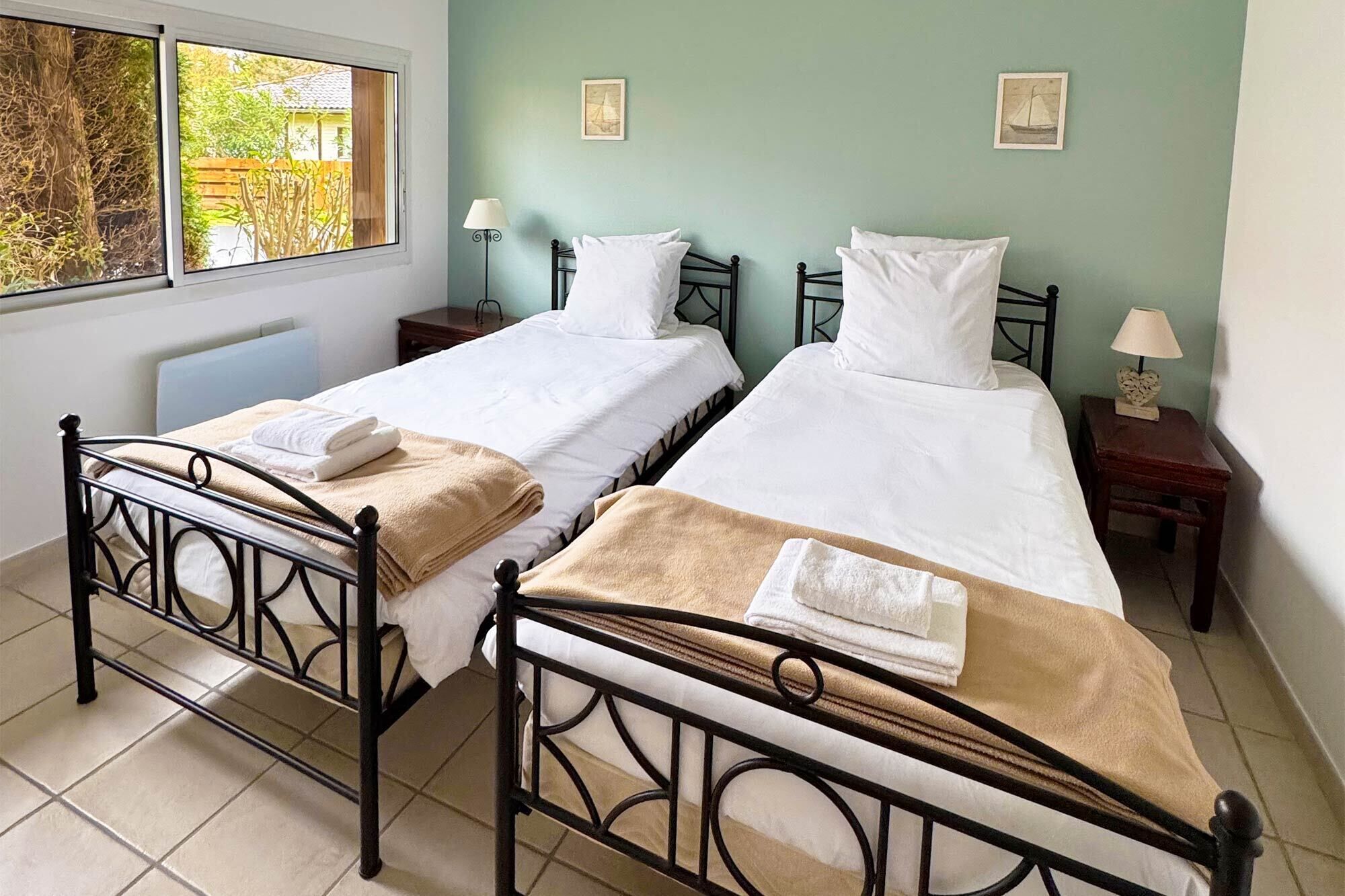 Two single beds with metal frames, white bedding, and towels, in a room with a view of greenery through a window.